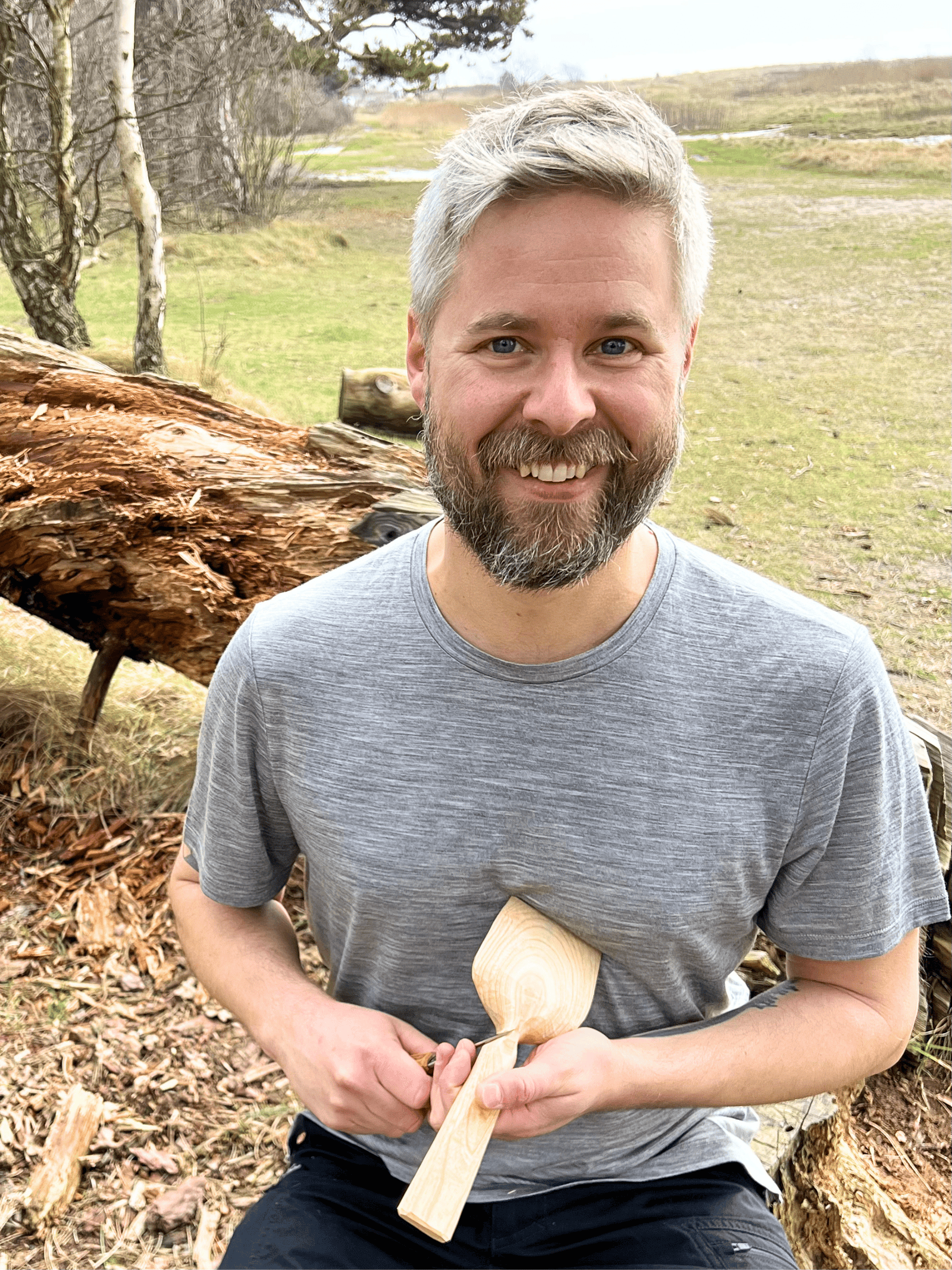 Sebastian Ungh outdoors with woodworking tools Male woodworker smiling with a wooden spatula in a natural outdoor setting.